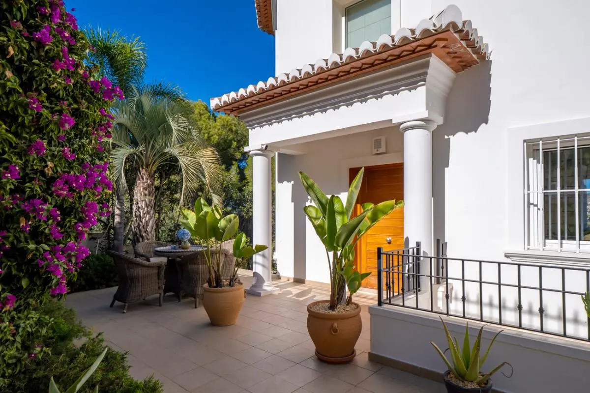 Main entrance featuring a white pillared porch, wooden door, and tiled terrace with wicker seating and lush Mediterranean landscaping.