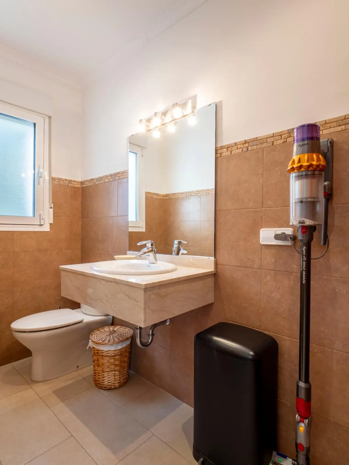 Modern bathroom featuring a marble floating vanity, integrated sink, and tan wall tiling. Includes a wall-mounted vacuum and natural light from a frosted window.