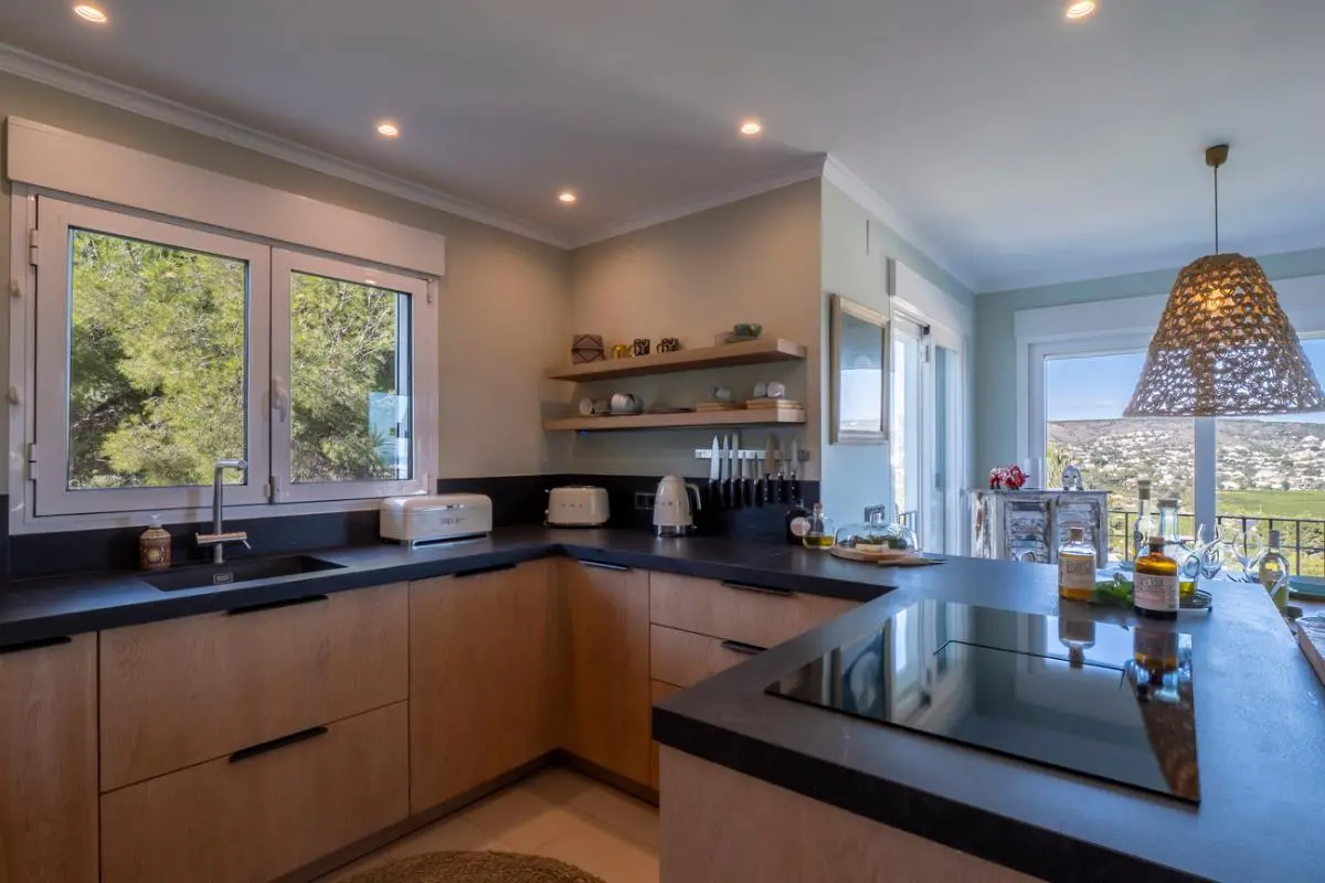 Modern kitchen featuring light wood cabinetry, dark countertops, and an induction hob. Large windows provide natural light and views of the surrounding pine trees and valley.