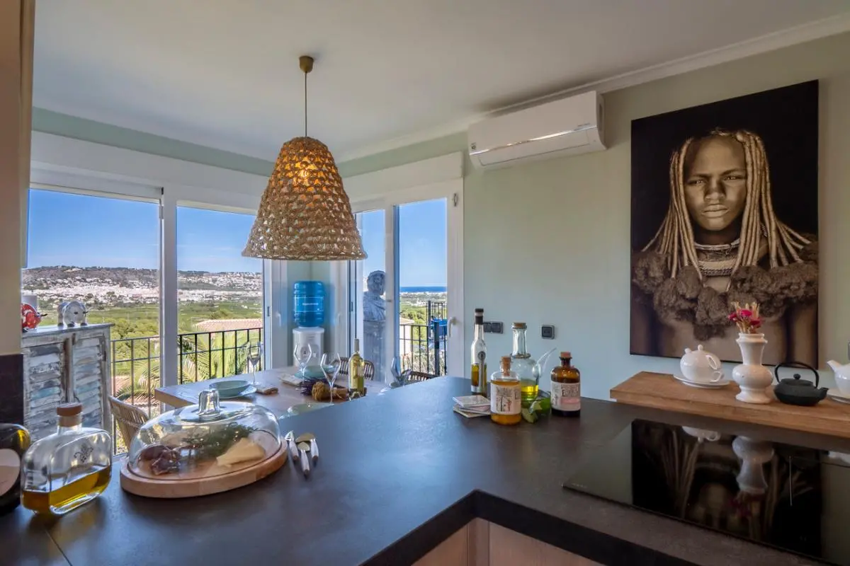 Modern kitchen with a dark countertop and induction hob, featuring a dining area with a woven pendant lamp and panoramic views of the coast through glass doors.