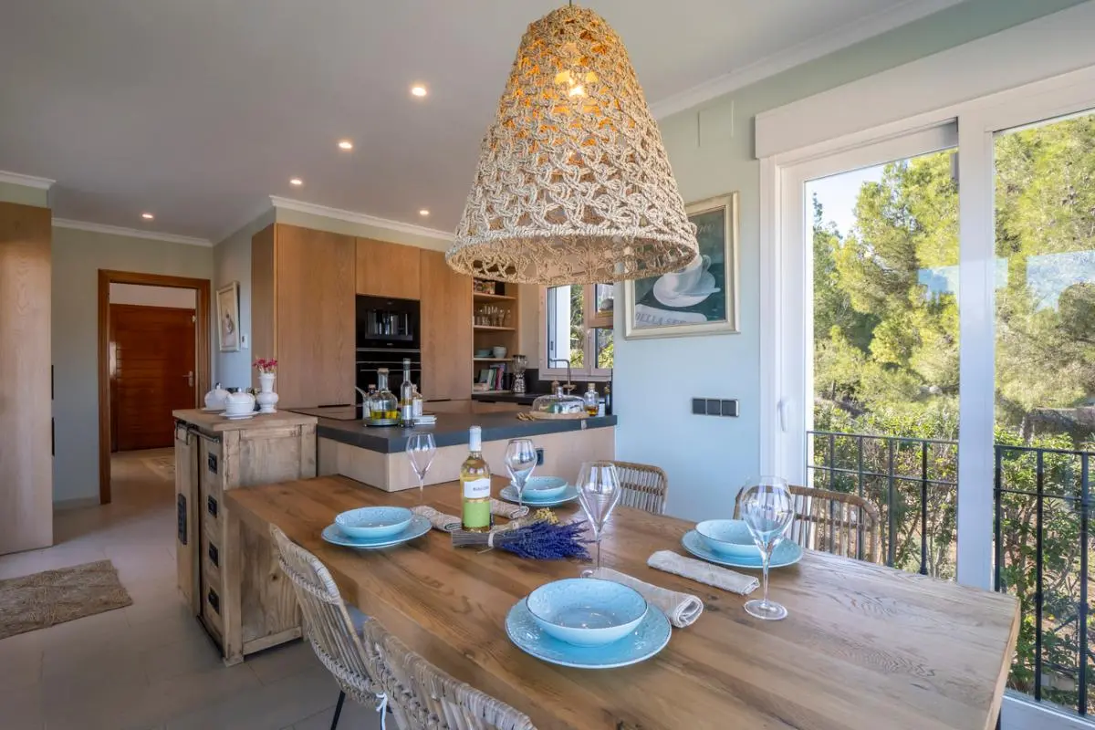 Open-plan dining area featuring a solid wood table, woven pendant light, and integrated kitchen with oak cabinetry. Large sliding doors offer views of the garden.