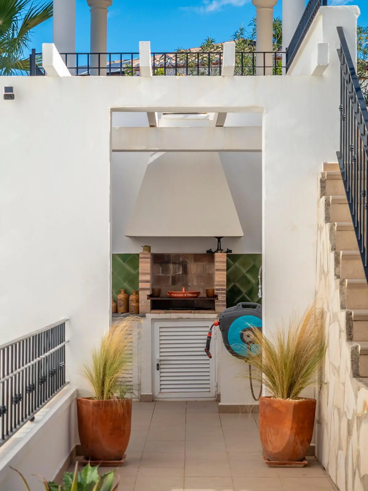 Outdoor built-in barbecue area featuring a brick grill, white chimney hood, and green tiled backsplash, framed by terracotta planters and white architectural walls.