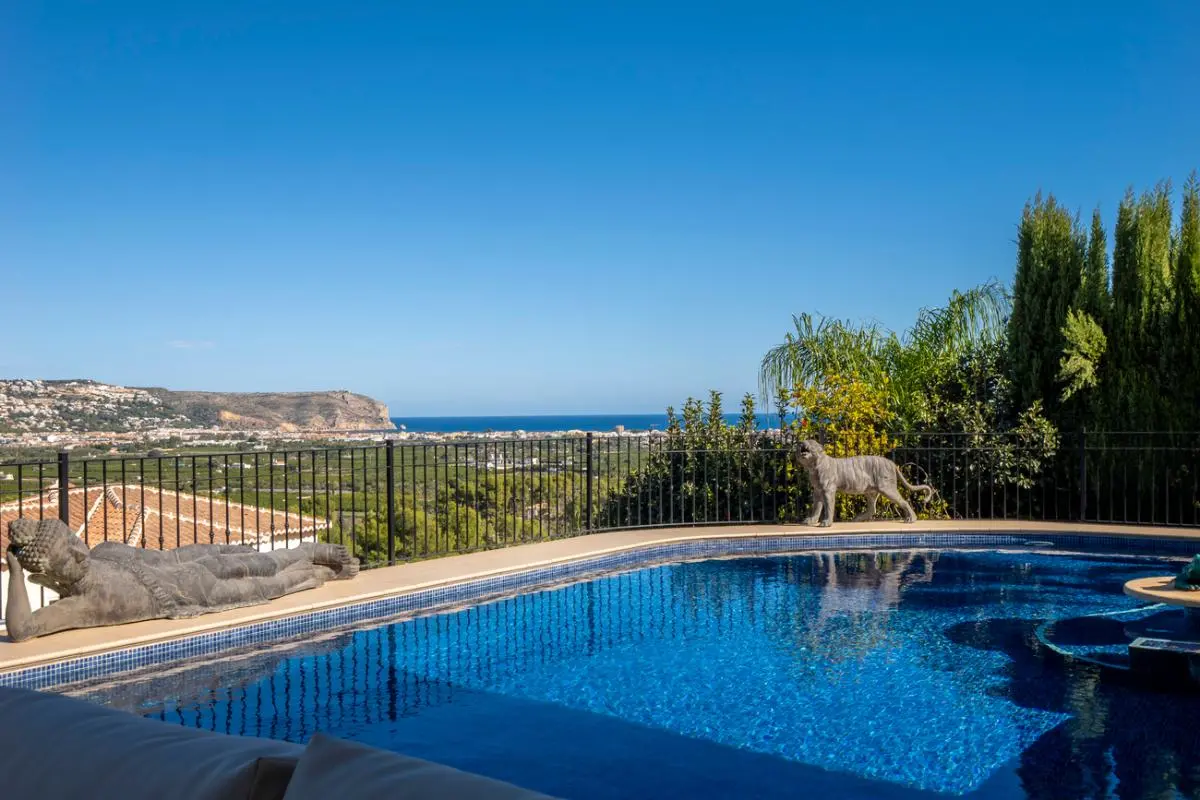 Outdoor swimming pool with blue mosaic tiles, featuring stone statues and panoramic views of the Mediterranean Sea and coastal landscape.
