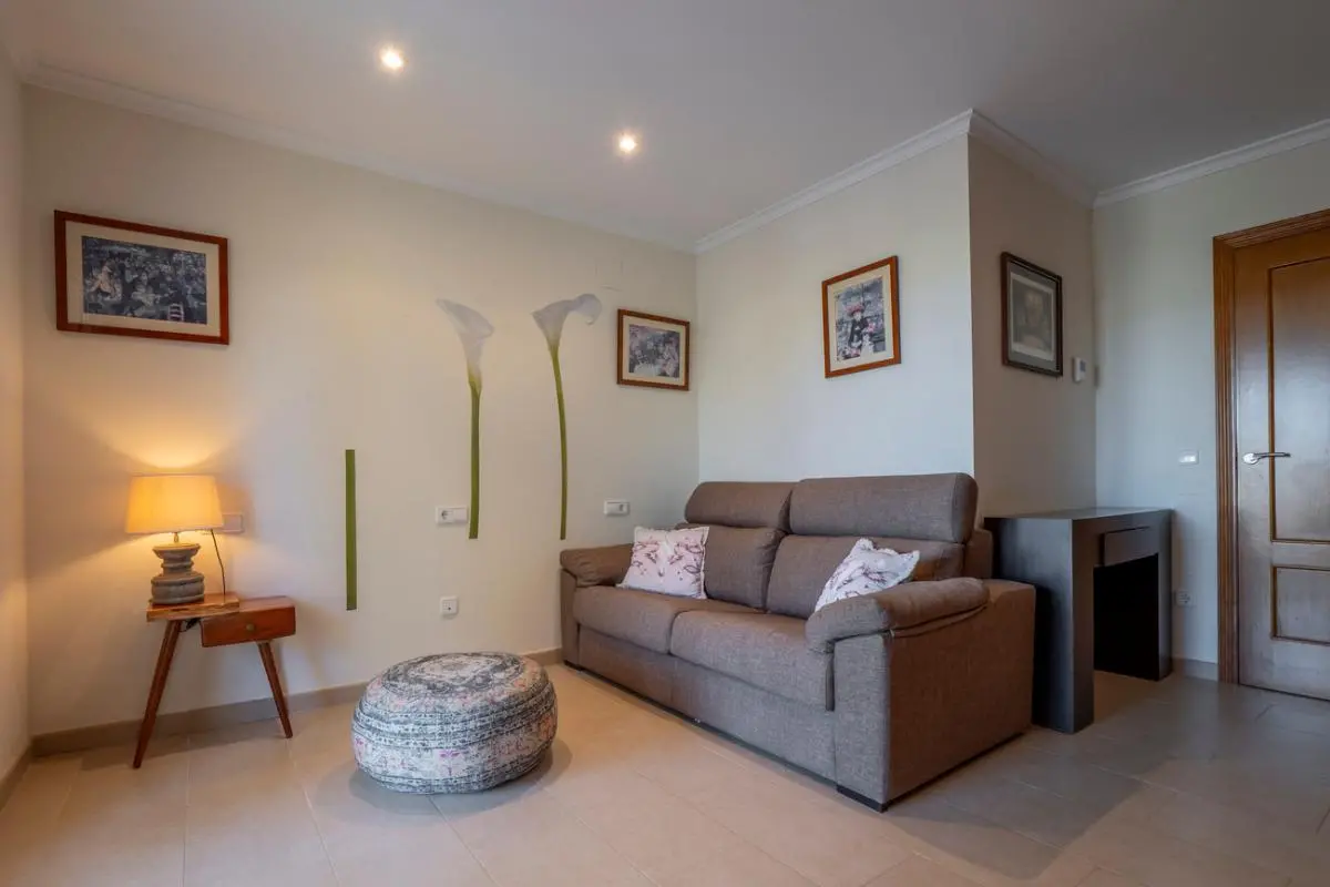Seating area featuring a brown fabric sofa, a patterned pouf, and a wooden side table. The wall is decorated with floral decals, framed art, and recessed lighting.