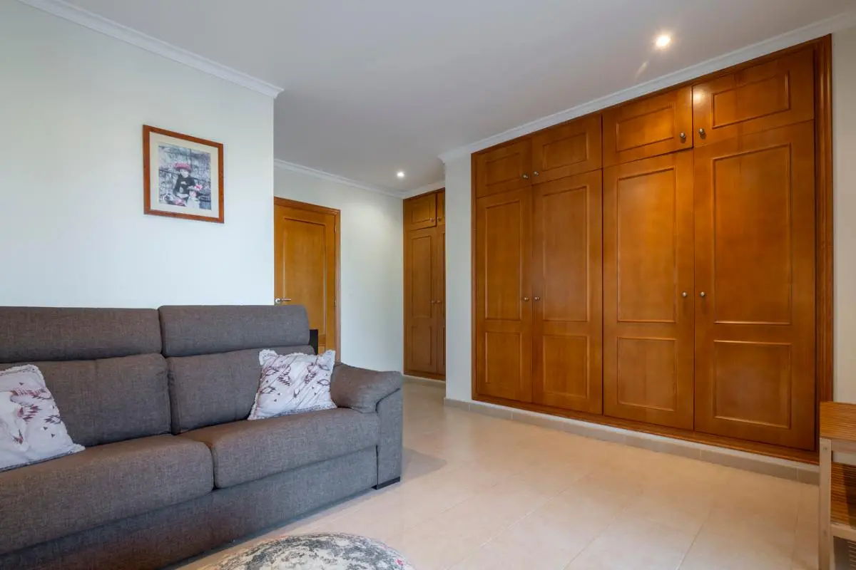 Spacious room featuring a large built-in wooden wardrobe with paneled doors, a grey fabric sofa, and light tiled flooring under recessed lighting.