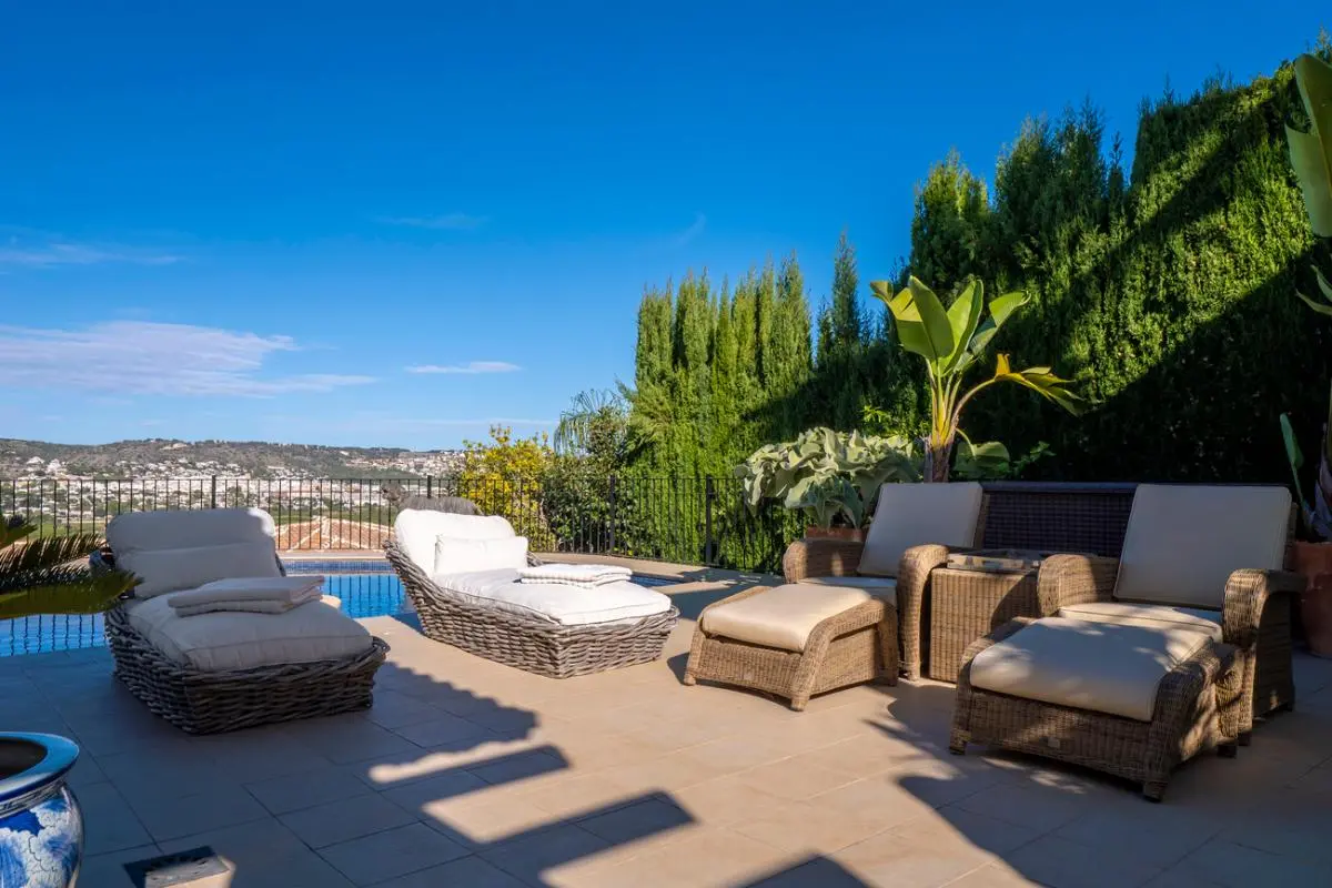 Terrace with wicker lounge chairs and white cushions beside a pool, offering panoramic hill views and surrounded by cypress trees.