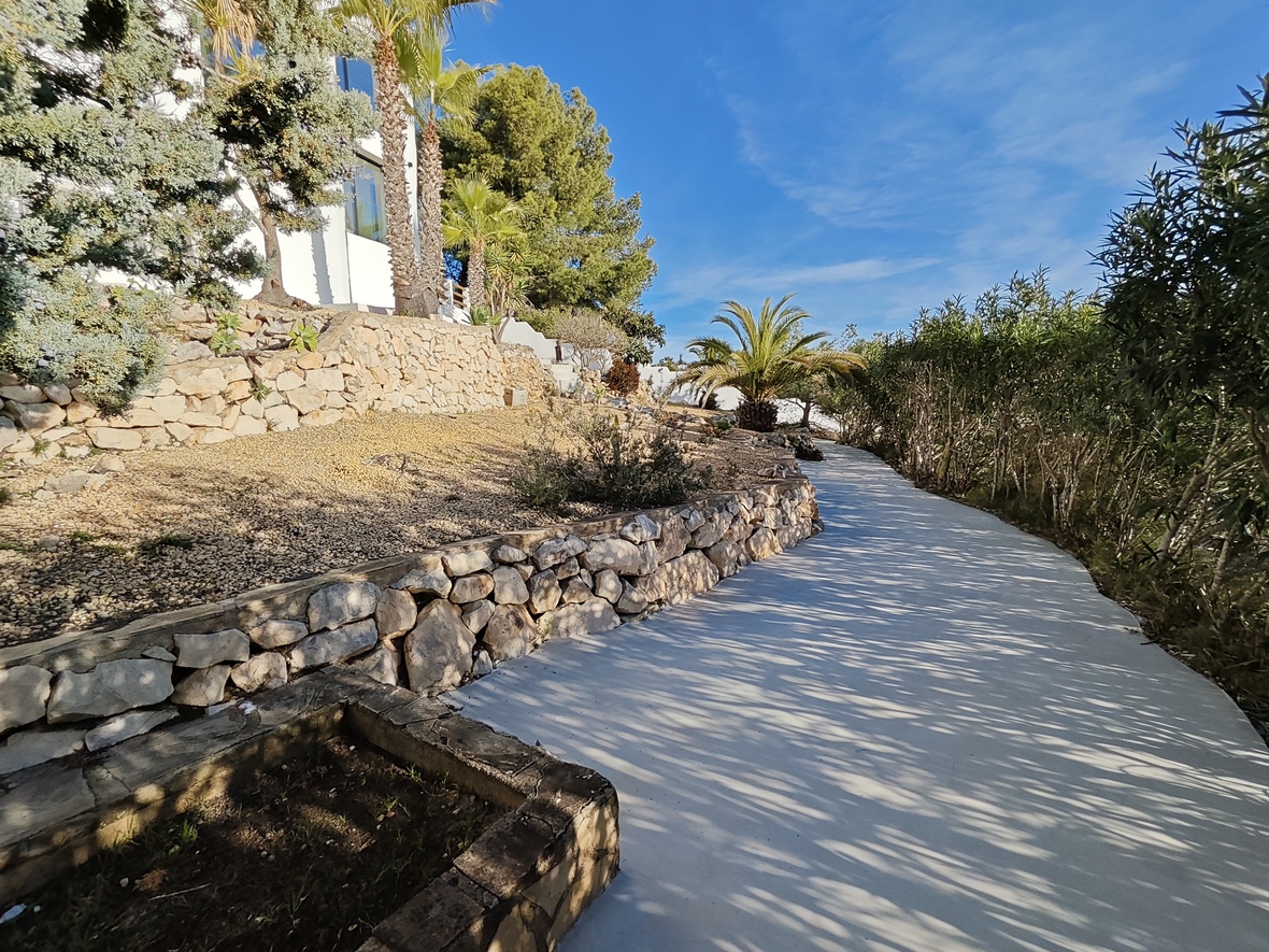 A curved paved pathway winds through a landscaped Mediterranean garden featuring dry stone retaining walls, palm trees, and mature pines.