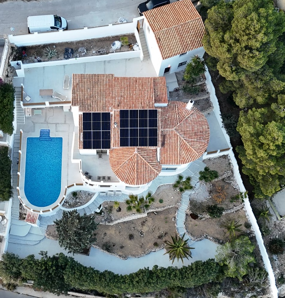 Aerial view of a Mediterranean villa featuring solar panels on a terracotta roof, a curved swimming pool, and terraced gardens with native vegetation.