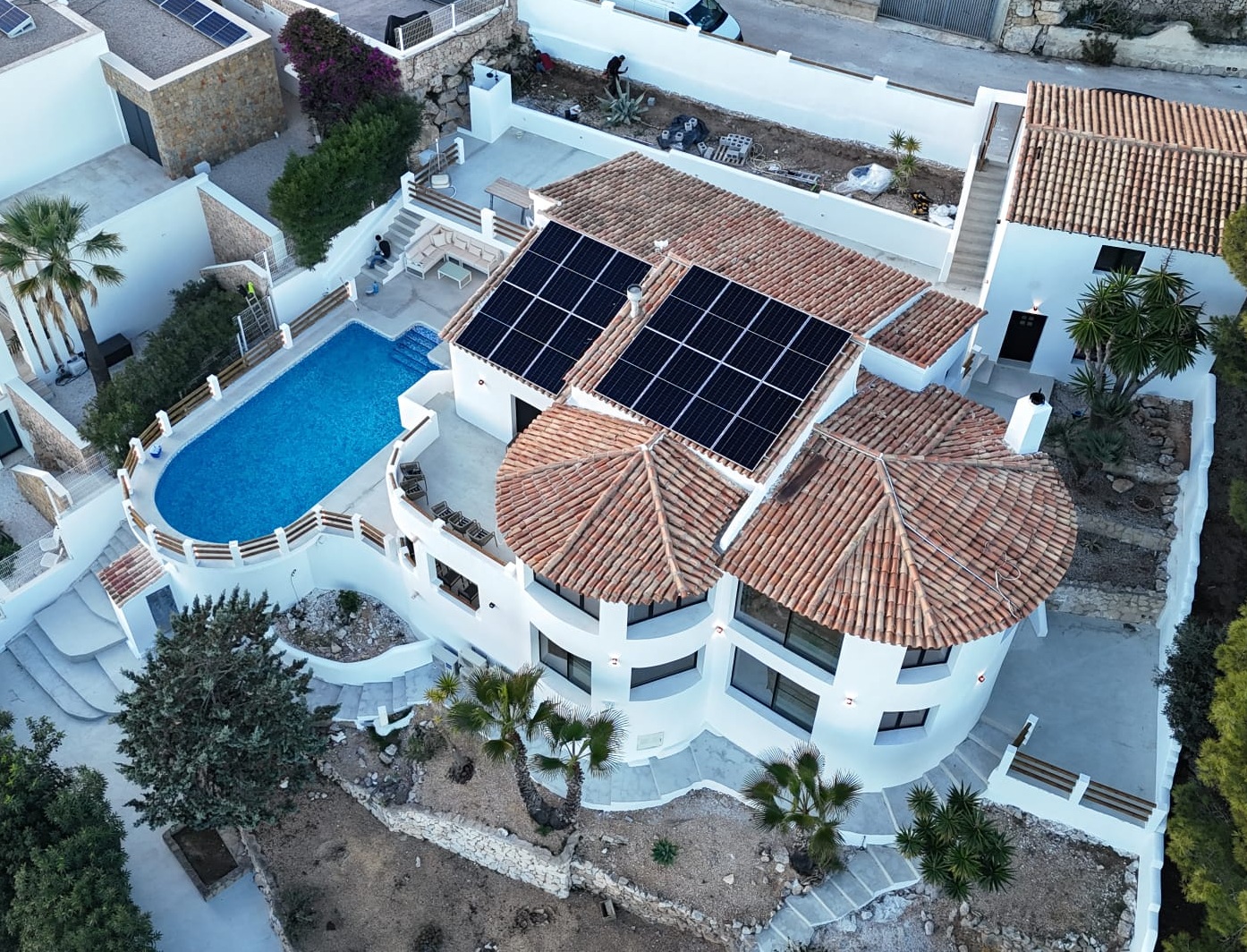 Aerial view of a multi-level Mediterranean villa featuring terracotta roofs with solar panels, an oval pool, and curved architectural lines set among stone terraces.