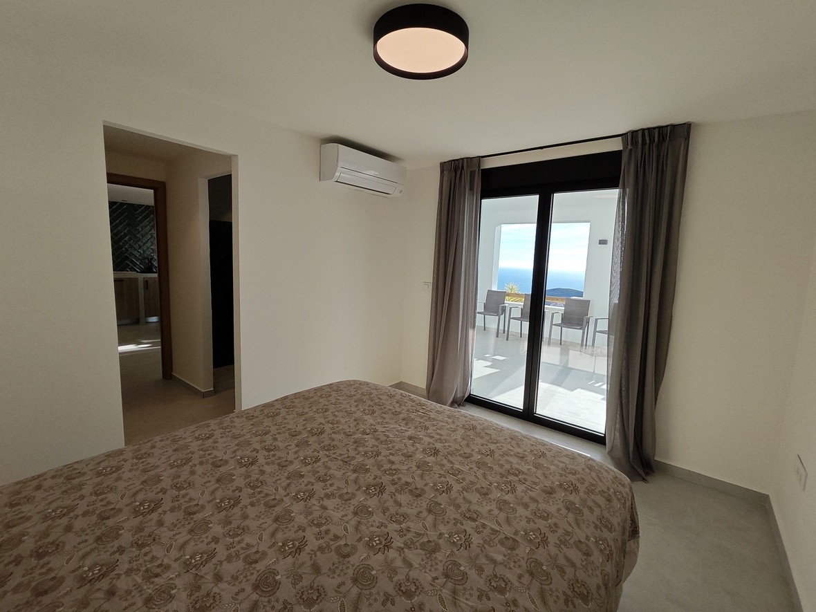 Bedroom with direct access to a terrace overlooking the sea. Features an air conditioning unit, neutral decor, and large sliding glass doors with grey curtains.
