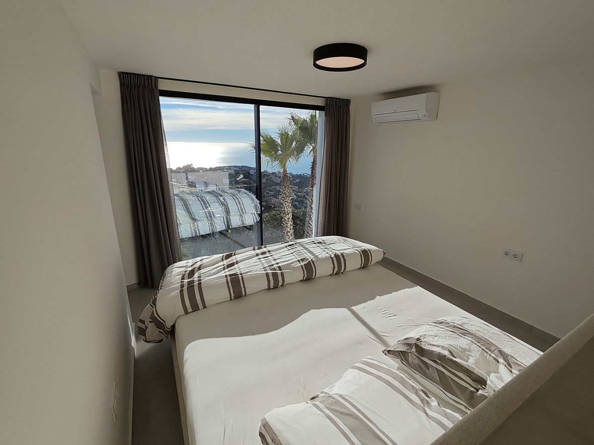 Bedroom with floor-to-ceiling sliding glass doors offering sea views and palm trees. Features minimalist white walls, striped bedding, and wall-mounted air conditioning.