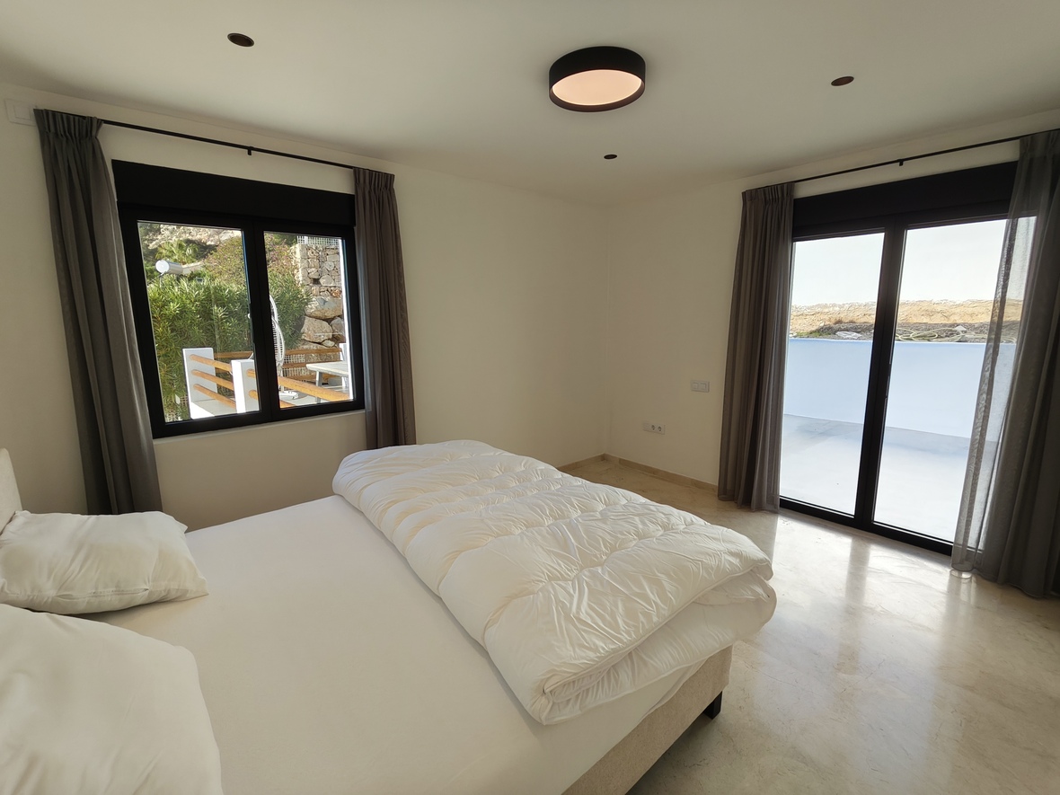 Bright bedroom featuring marble flooring, black-framed sliding doors to a terrace, and a minimalist ceiling light. Grey curtains complement the neutral walls.