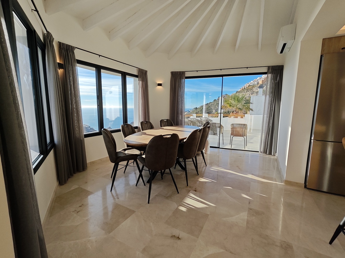 Bright dining area with an oval wooden table, marble flooring, and a vaulted beamed ceiling. Large windows provide panoramic sea views and direct terrace access.