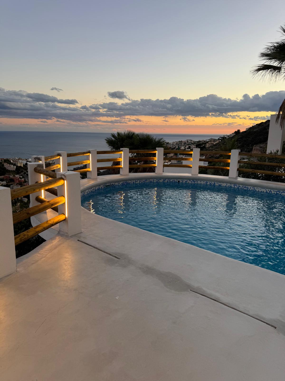 Circular swimming pool with blue mosaic tiling and integrated LED lighting in the wooden railing, overlooking the Mediterranean coast at sunset.