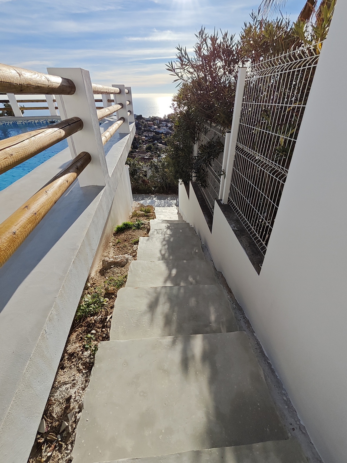 Concrete exterior staircase with white walls and wooden railings, offering views of the pool and the Mediterranean coastline under bright natural light.