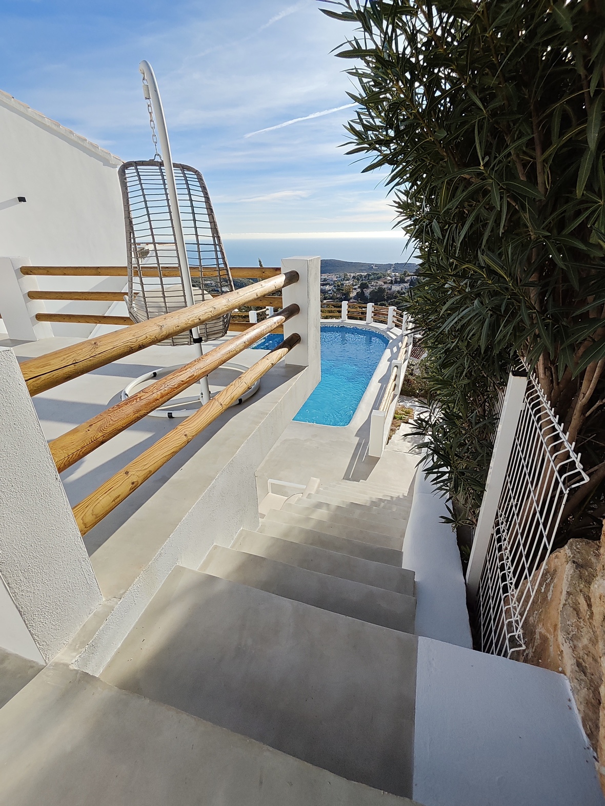 Concrete stairs lead to a terrace with a hanging wicker chair and wooden railings, overlooking a swimming pool and the Mediterranean coastline.