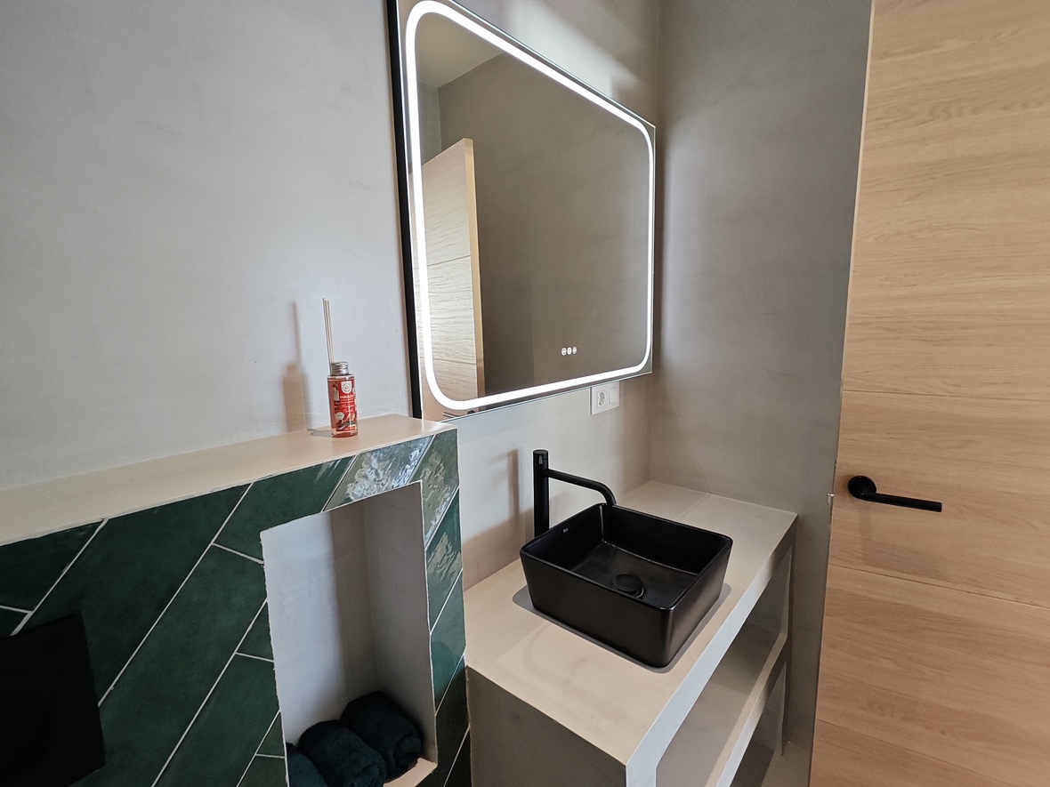 Contemporary bathroom with a black vessel sink, integrated LED mirror, and emerald green herringbone tile accents against microcement walls.