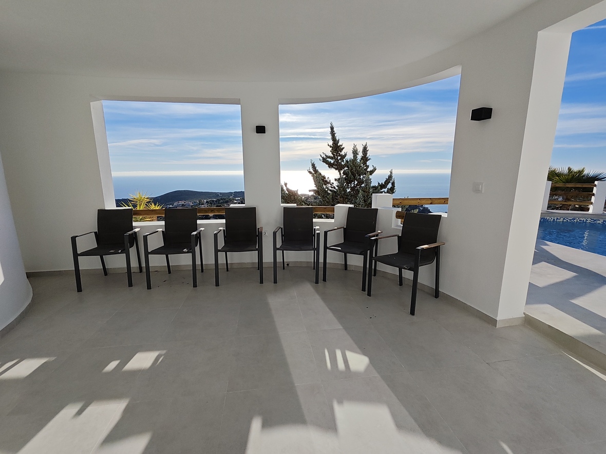 Covered terrace featuring modern black armchairs, light grey tiling, and large wall openings framing panoramic views of the sea and the adjacent swimming pool.