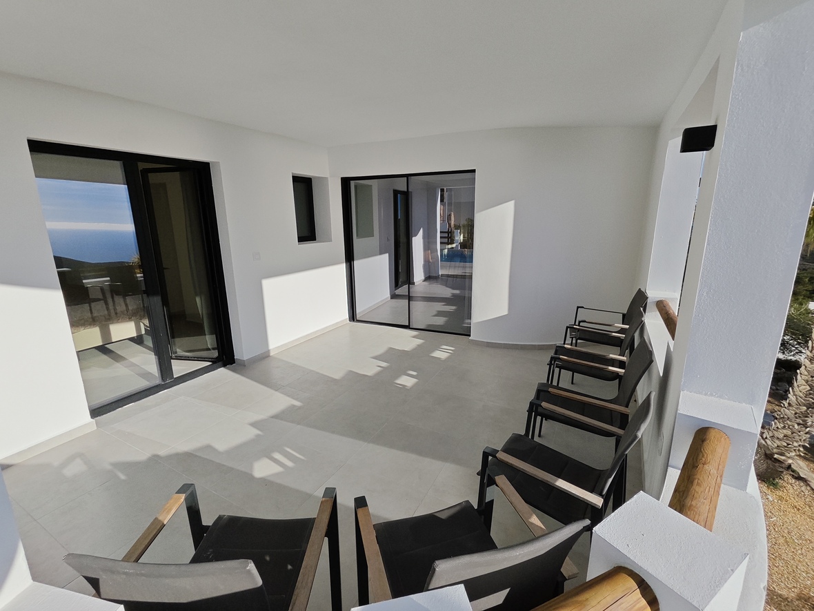 Covered terrace with white walls, light grey tiling, and black-framed glass doors. Features modern black seating with wooden armrests and sea views.