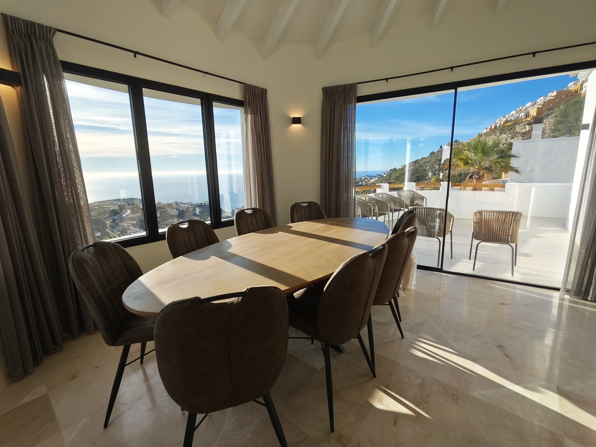 Dining area featuring an oval wooden table and marble floors, offering panoramic sea views through large windows and access to a sun-drenched terrace.