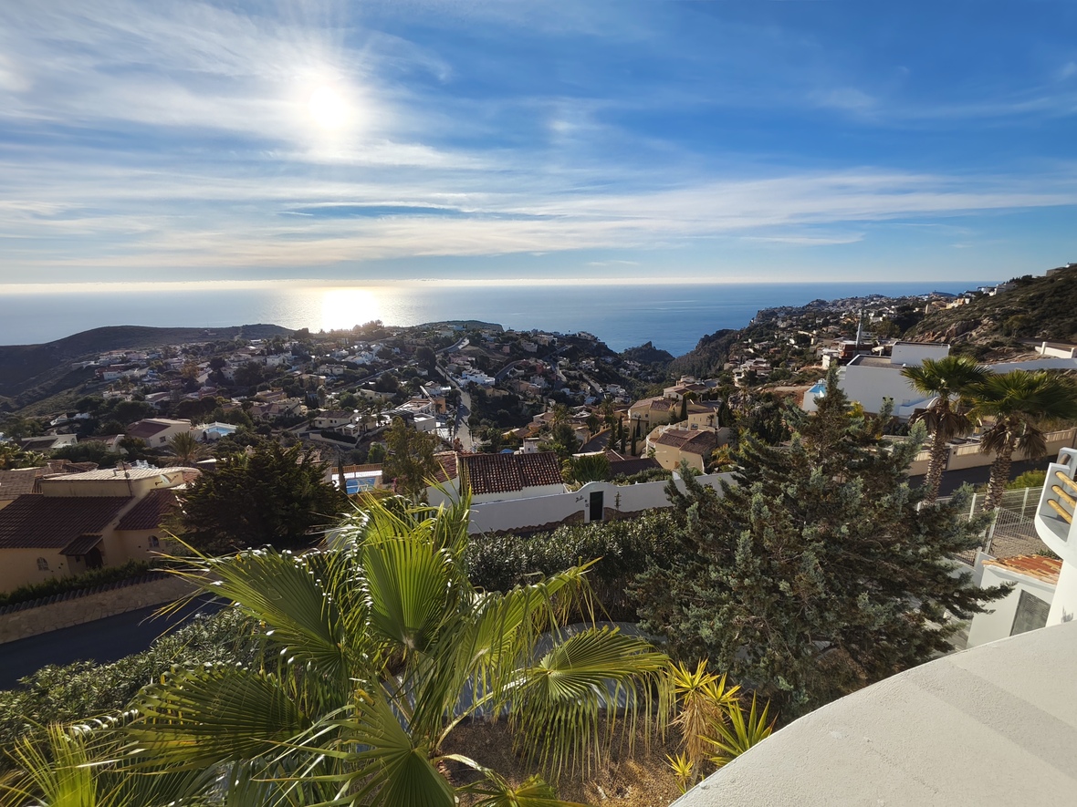 Elevated panoramic view overlooking a coastal residential area and the Mediterranean Sea, featuring white villas, terracotta roofs, and lush palm trees.