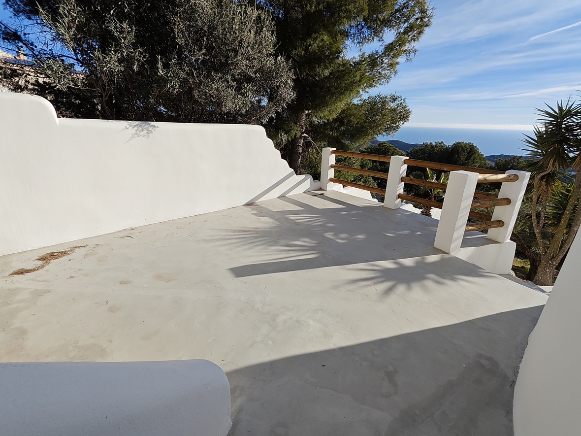 Elevated terrace with smooth concrete flooring, white plastered walls, and a wooden railing offering panoramic views of the Mediterranean Sea and pine forest.