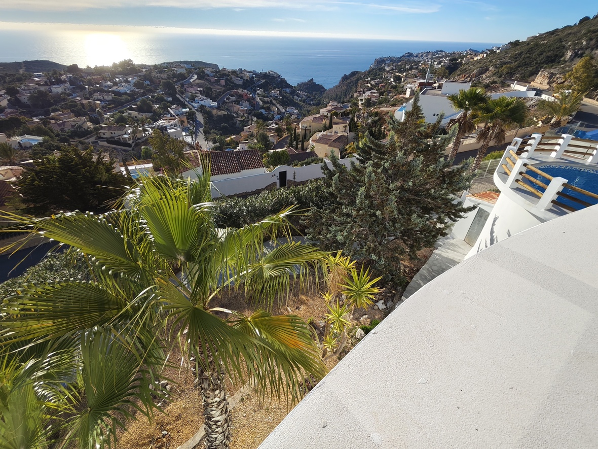 Elevated view from a white terrace overlooking a circular pool with wooden railings, Mediterranean gardens, and panoramic sea views.