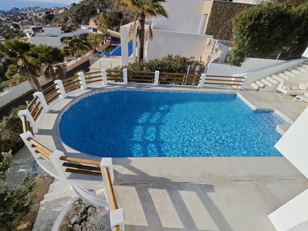 Elevated view of a curved outdoor pool with blue mosaic tiling, featuring a white and wood railing and panoramic views of the Mediterranean coastline.