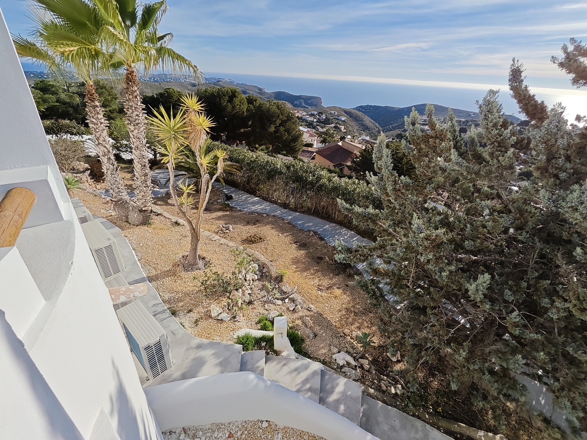 Elevated view of a landscaped garden featuring palm trees and gravel paths, overlooking a coastal valley with panoramic Mediterranean Sea views.