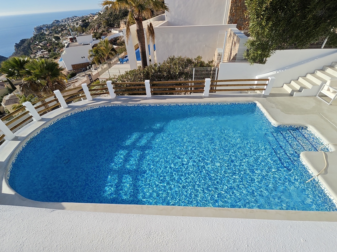 Elevated view of a private mosaic-tiled pool with integrated steps, featuring a white terrace and wooden railing overlooking the Mediterranean coastline.