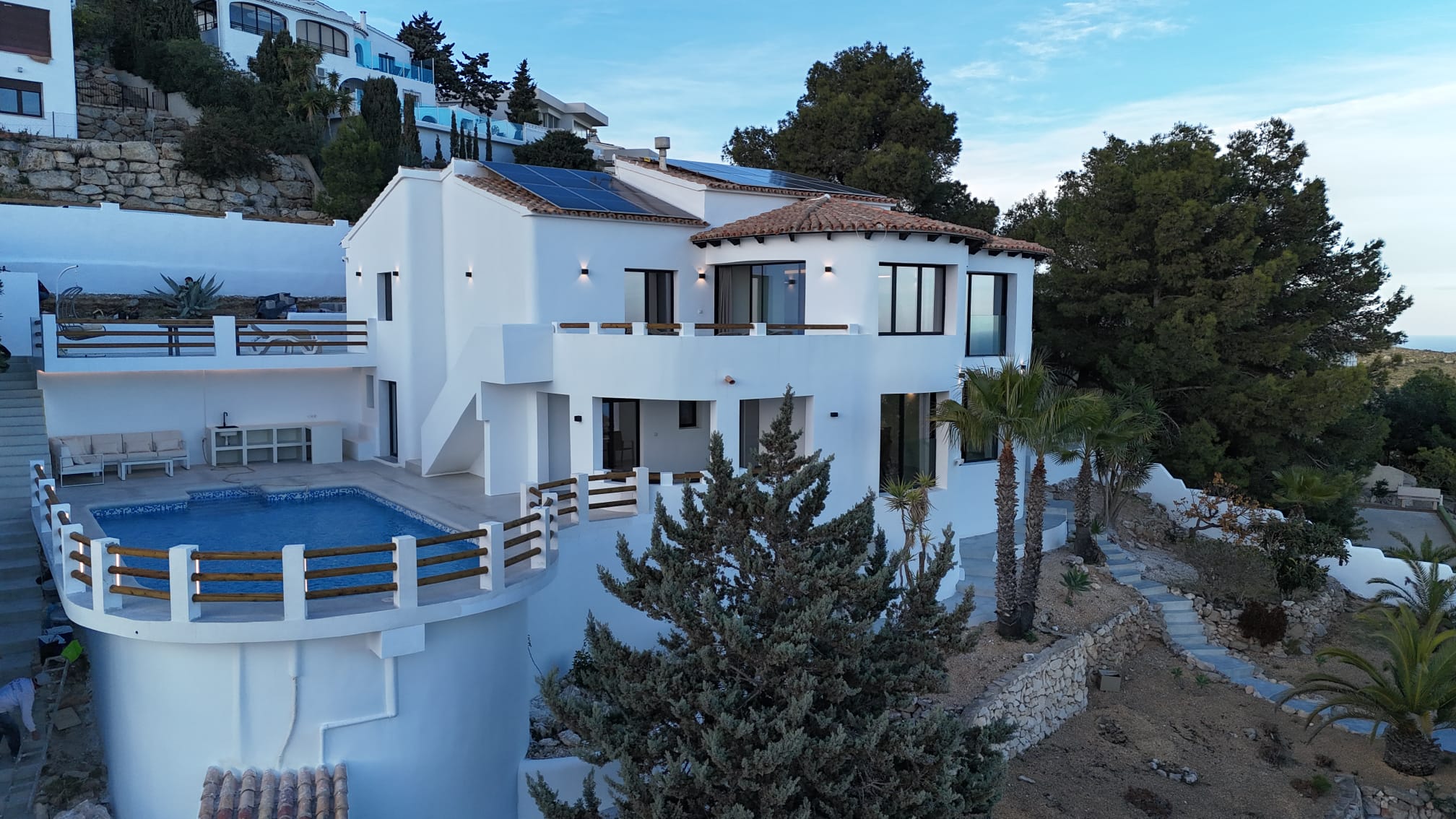 Exterior of a multi-level white villa featuring a curved swimming pool, solar panels on the roof, and terraced gardens with Mediterranean vegetation.