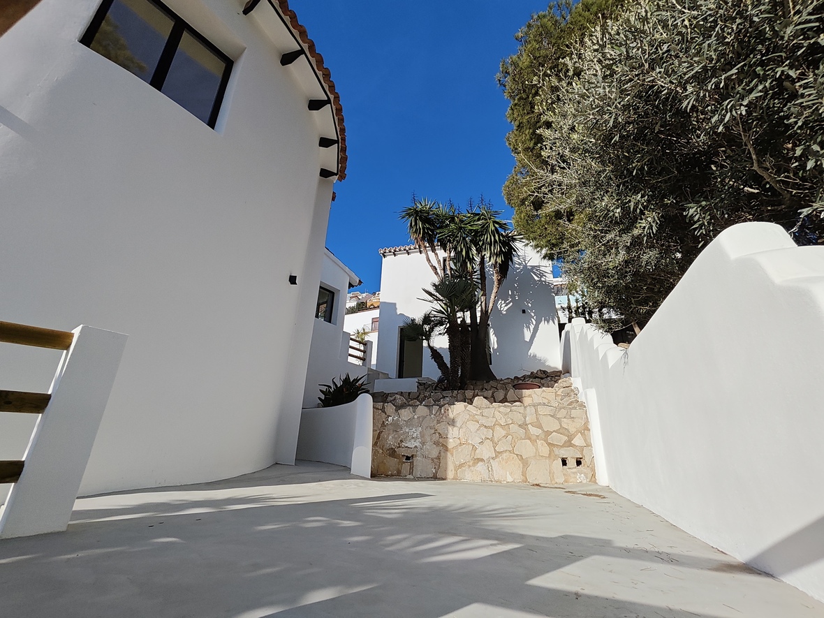Exterior view featuring white curved walls, a natural stone retaining wall, and paved grounds. Mediterranean landscaping includes palm trees and mature trees under a clear sky.