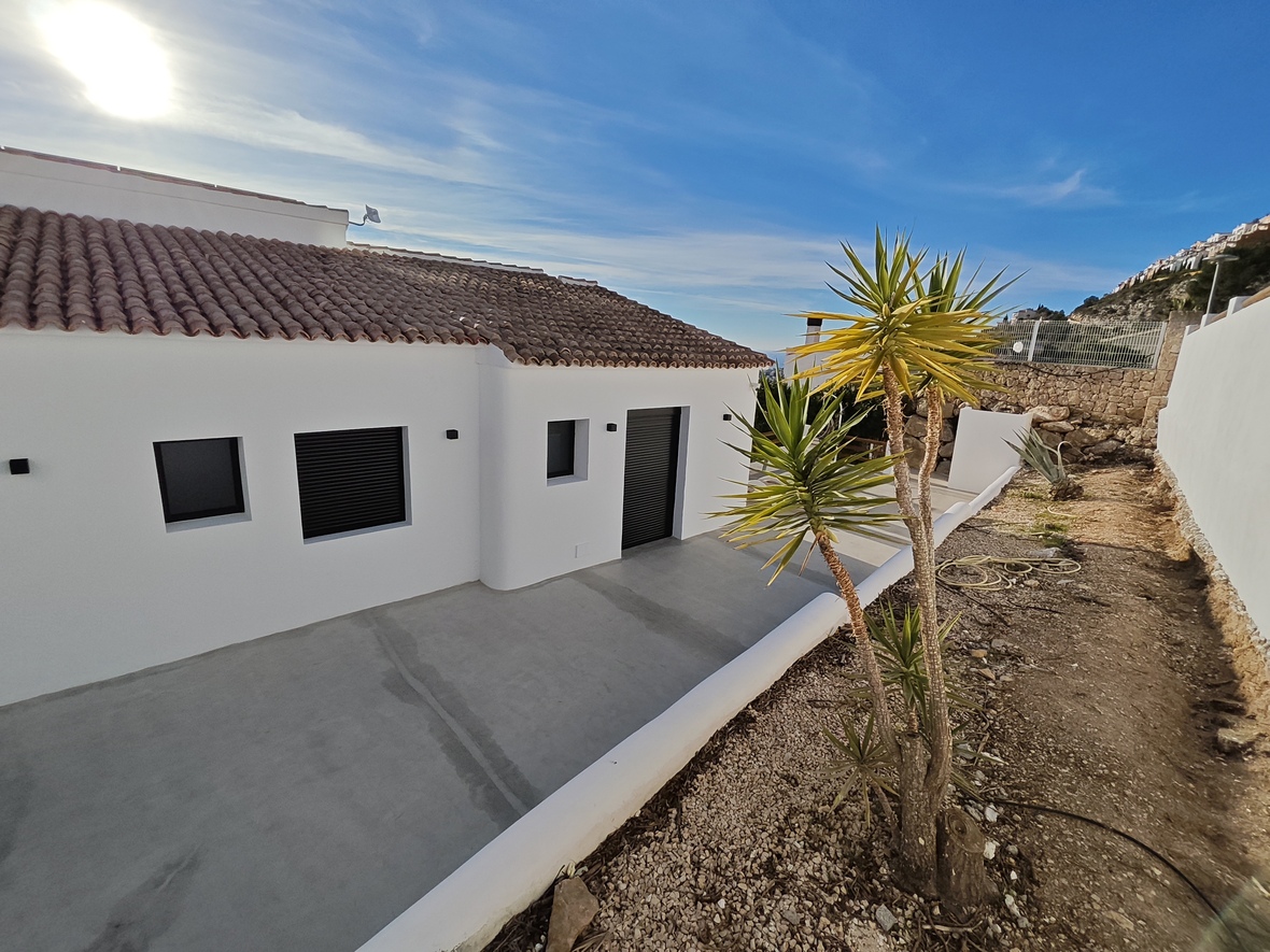 Exterior view of a minimalist white villa featuring a terracotta tile roof, dark shutters, and a concrete terrace bordered by a yucca plant and stone wall.
