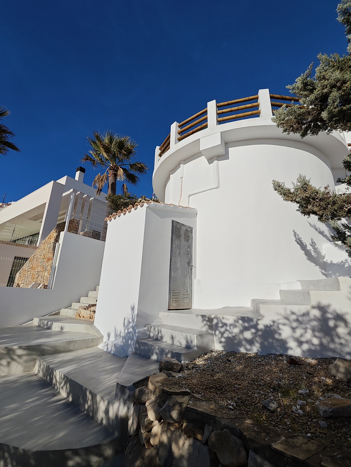 Exterior view of a white cylindrical structure featuring a rooftop terrace with wooden railings and integrated concrete stairs under a clear blue sky.