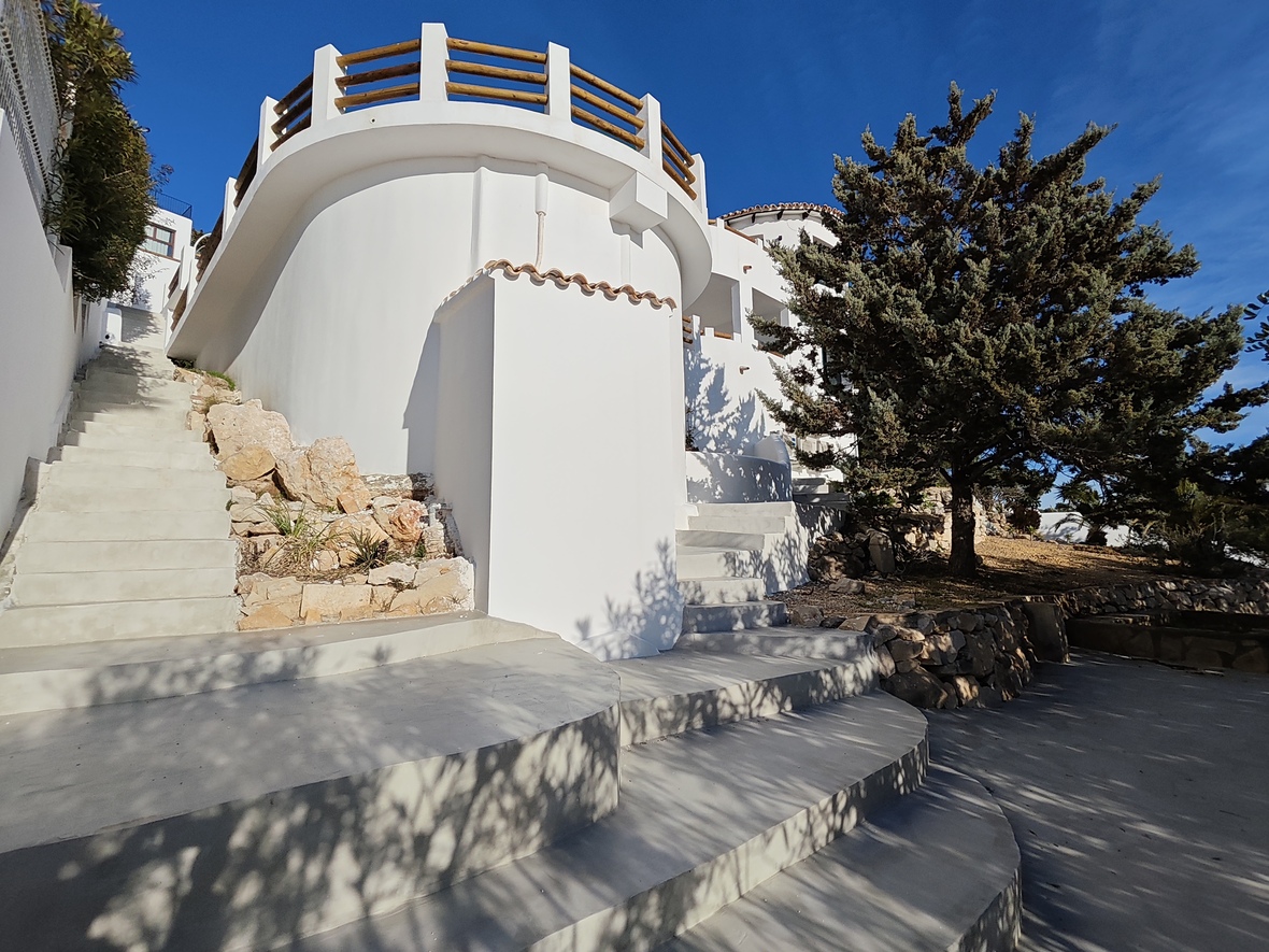 Exterior view of a white Mediterranean villa featuring curved walls, wide concrete stairs, and a circular terrace with a wooden railing.
