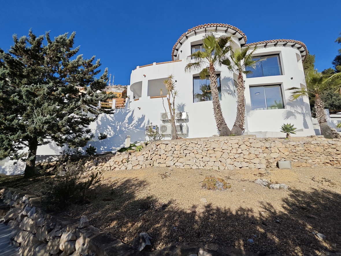 Exterior view of a white Mediterranean villa with curved walls, stone terraces, and palm trees against a clear blue sky.