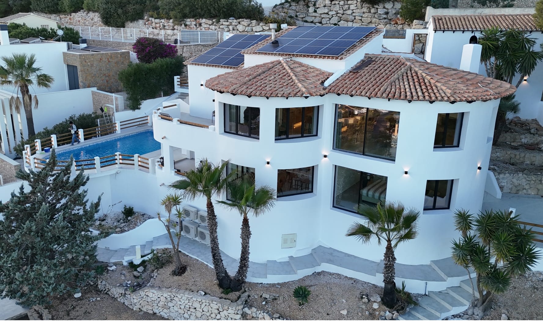 Exterior view of a white multi-level villa featuring curved architecture, terracotta tile roofs with solar panels, and a private pool area with palm trees.