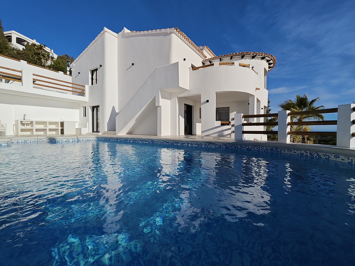 Exterior view of a white multi-level villa featuring curved terraces, wooden railings, and a blue mosaic-tiled swimming pool reflecting the architecture.
