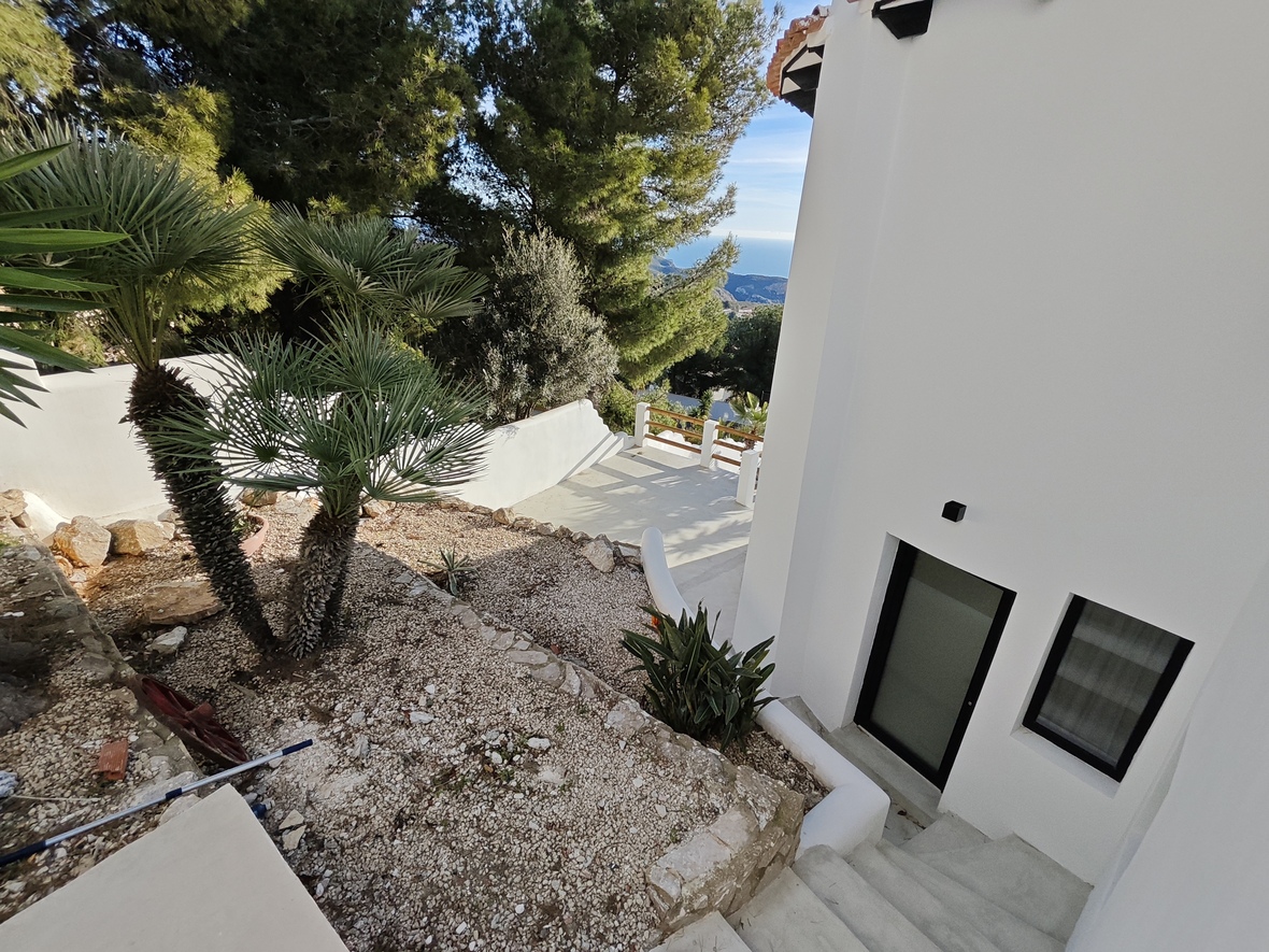 Exterior view of a white villa featuring a gravel garden with palms, stone walls, and concrete stairs leading to a black-framed entrance with sea views.