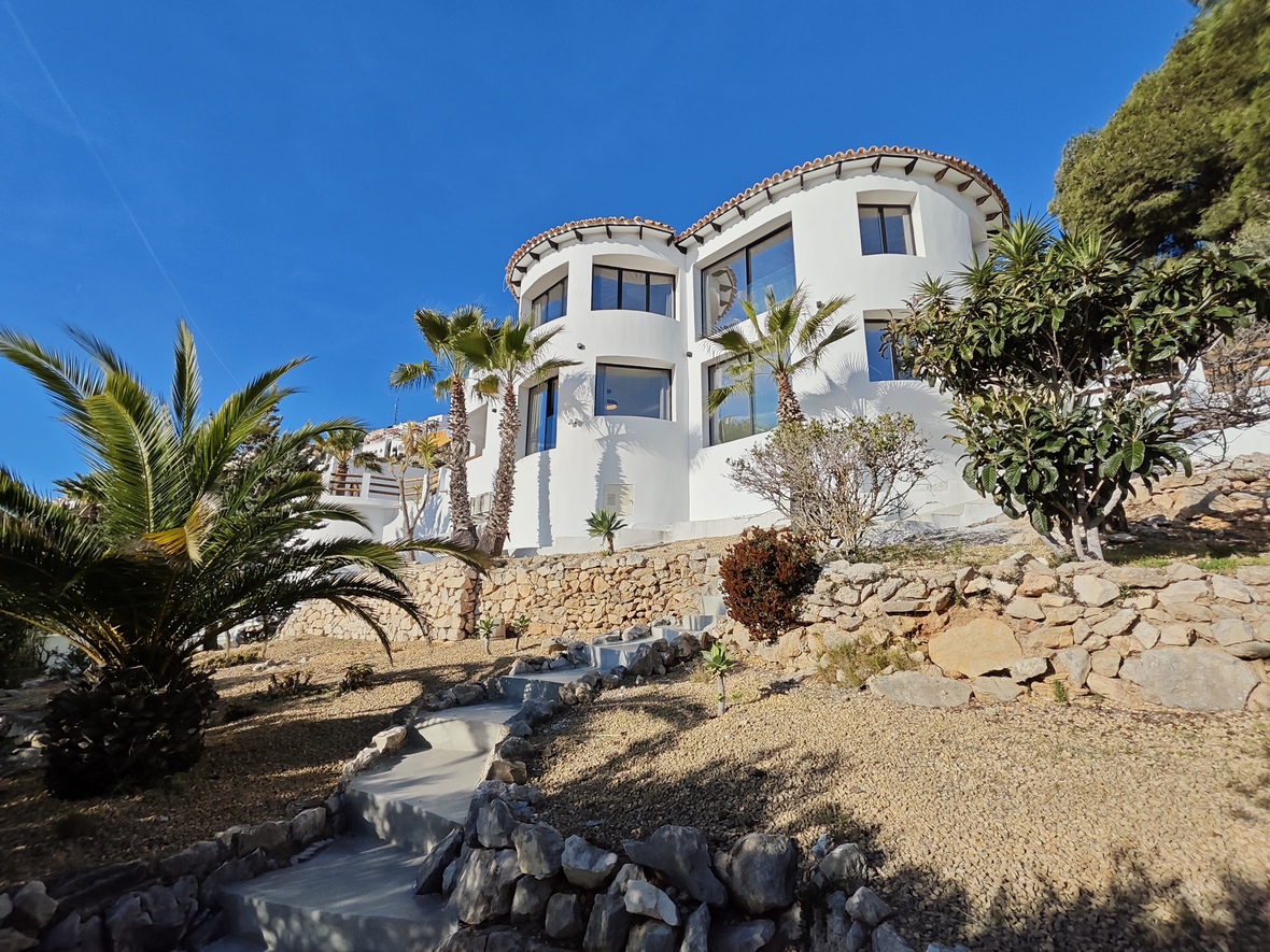 Exterior view of a white villa with curved architectural lines and large windows. The sloped garden includes stone retaining walls, palm trees, and a concrete staircase.