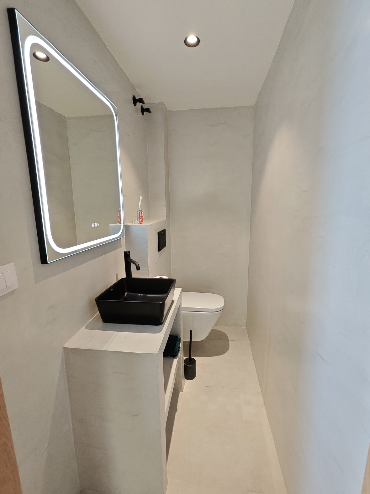 Minimalist bathroom featuring a black vessel sink, matte black faucet, backlit LED mirror, and seamless light gray microcement walls.