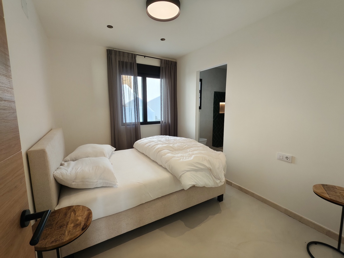 Minimalist bedroom featuring a beige upholstered bed, wooden side tables, and an en-suite bathroom with dark accent tiling. Natural light enters through black-framed windows.