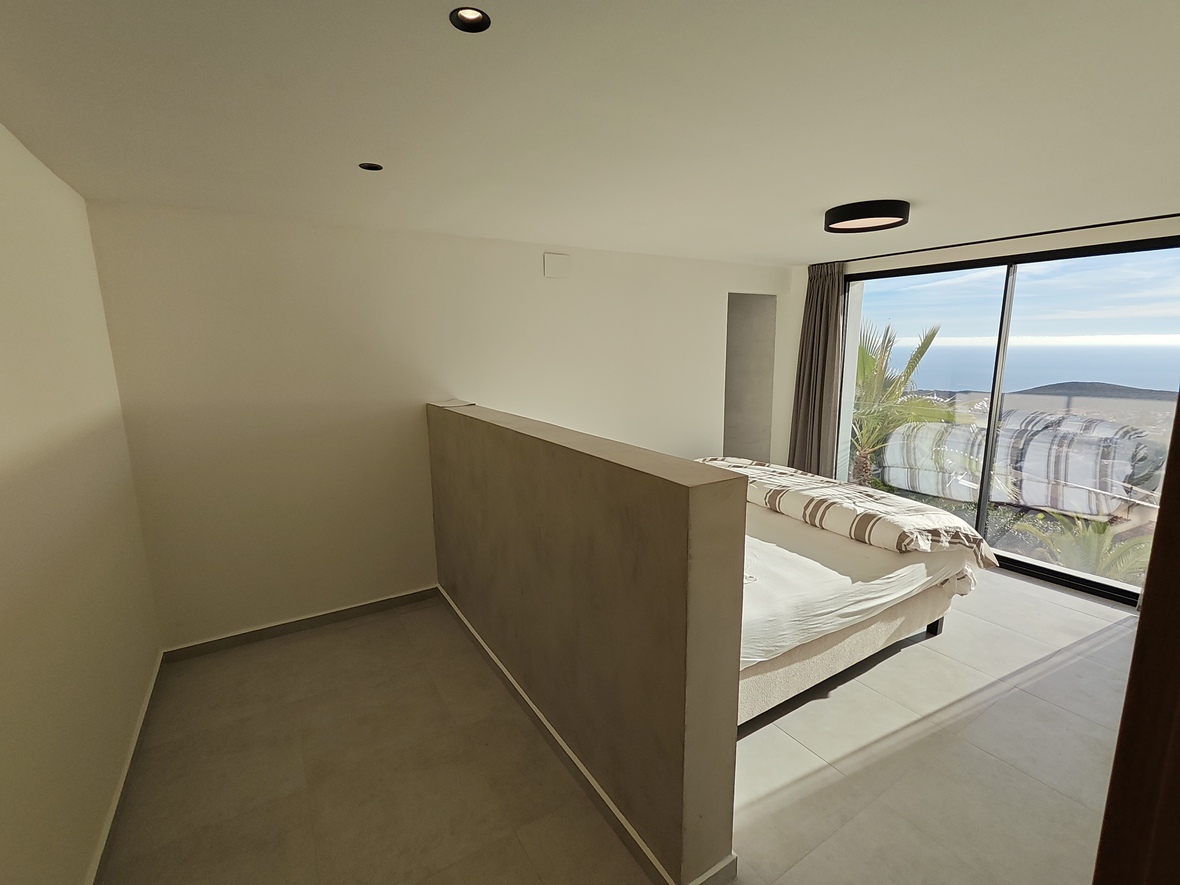 Minimalist bedroom featuring a low partition wall, large sliding glass doors with ocean views, and integrated recessed ceiling lighting.