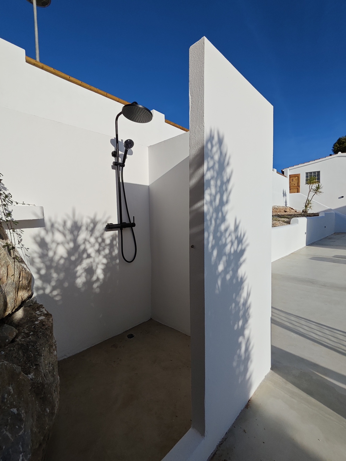 Minimalist outdoor shower featuring matte black fixtures against crisp white walls, set on a smooth concrete base under a clear blue sky.