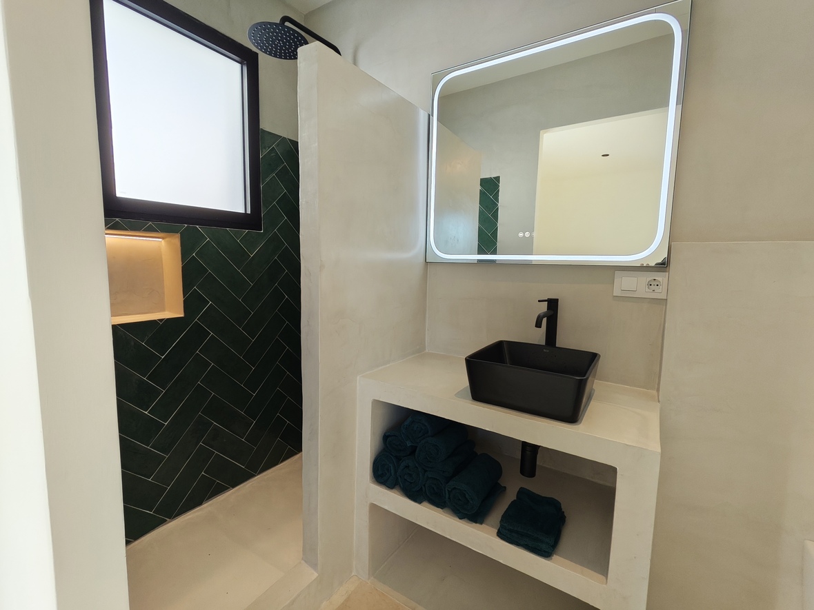 Modern bathroom featuring a black vessel sink on a microcement vanity, a backlit LED mirror, and a walk-in shower with emerald green herringbone tiles.