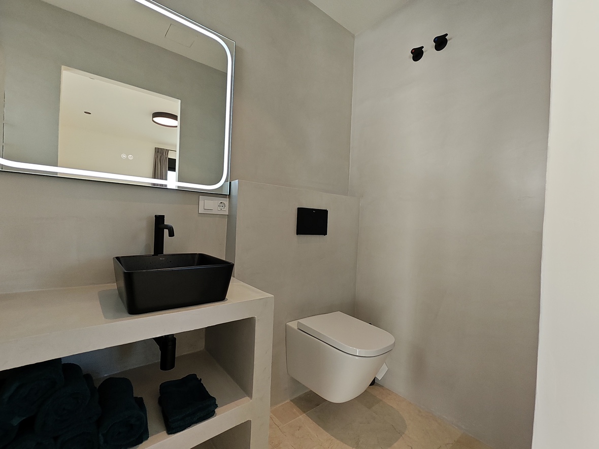 Modern bathroom featuring a black vessel sink on a microcement vanity, a backlit mirror, and a wall-mounted toilet against grey plaster walls.