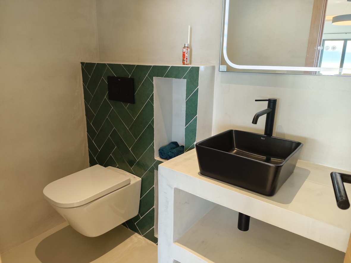 Modern bathroom featuring a black vessel sink on a microcement vanity, complemented by a green herringbone tile accent wall and a wall-hung toilet.