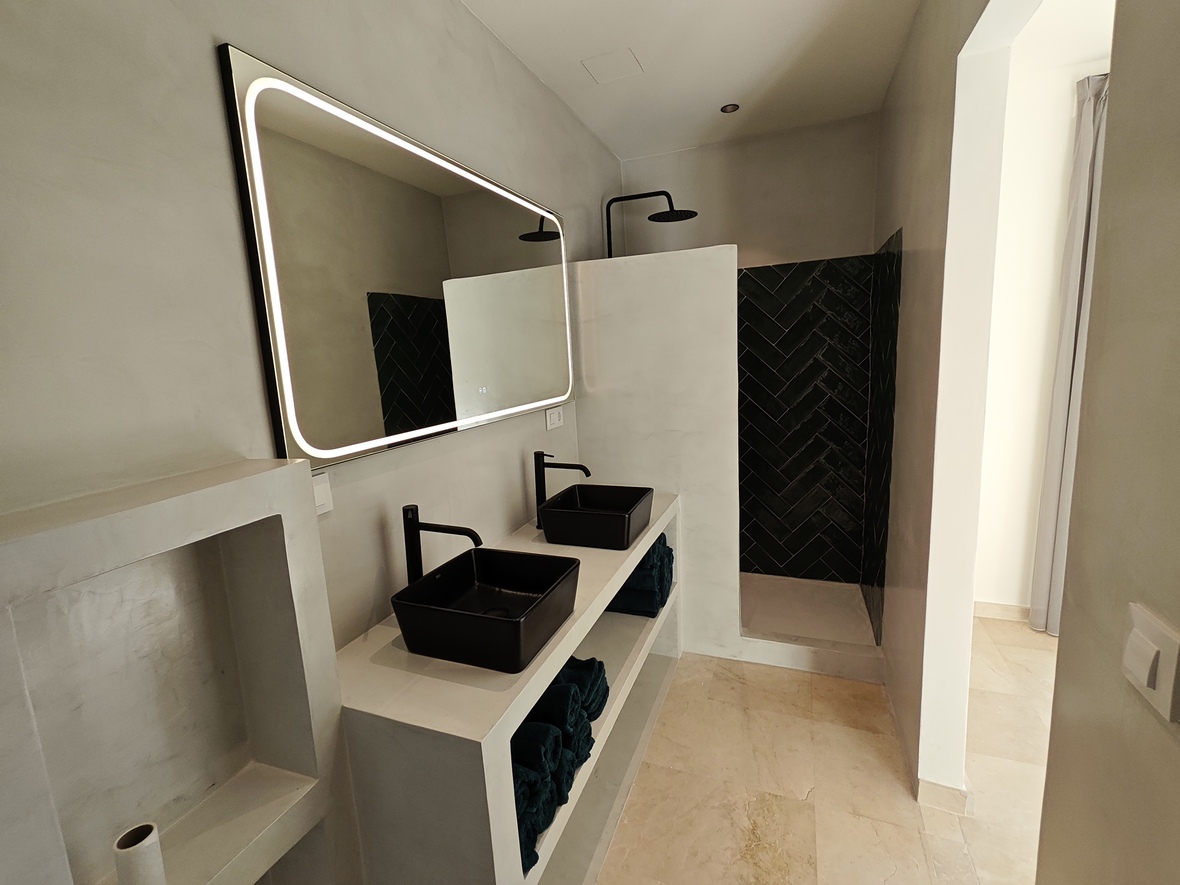 Modern bathroom featuring a microcement double vanity with black vessel sinks, a large backlit mirror, and a walk-in shower with black herringbone tiling.