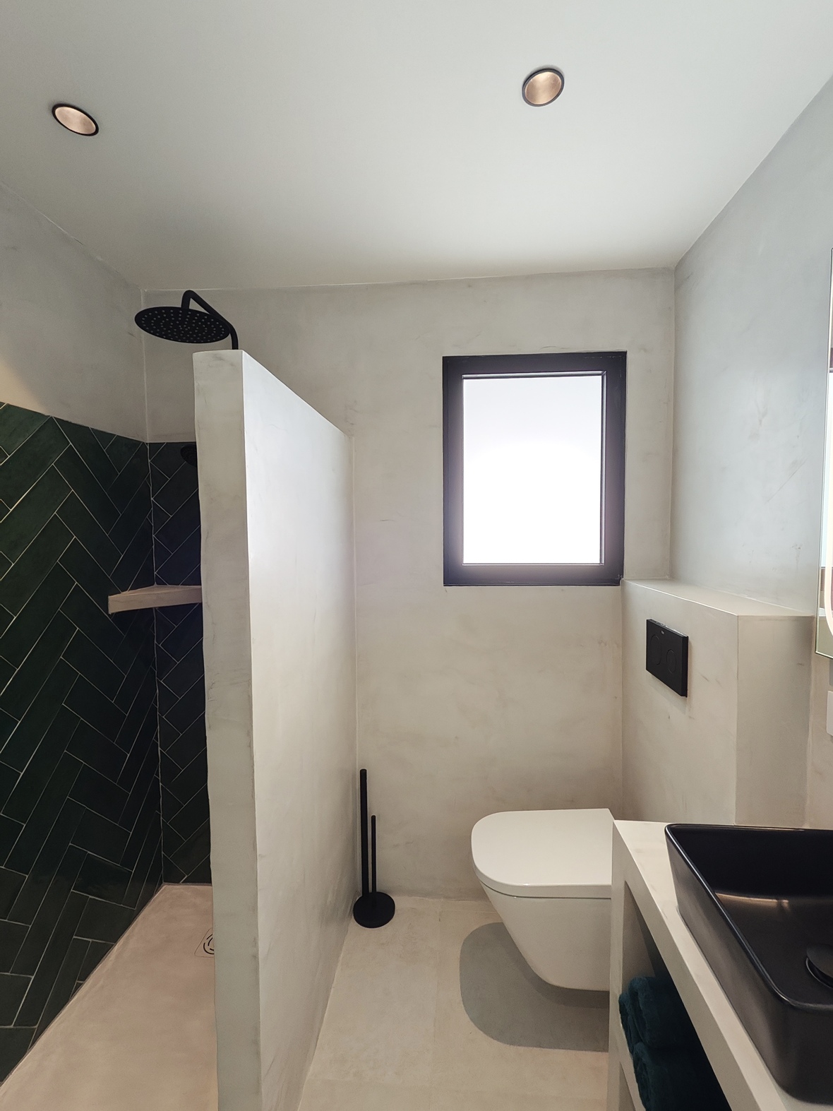 Modern bathroom featuring a walk-in shower with green herringbone tiles, a wall-mounted toilet, and white microcement finishes.