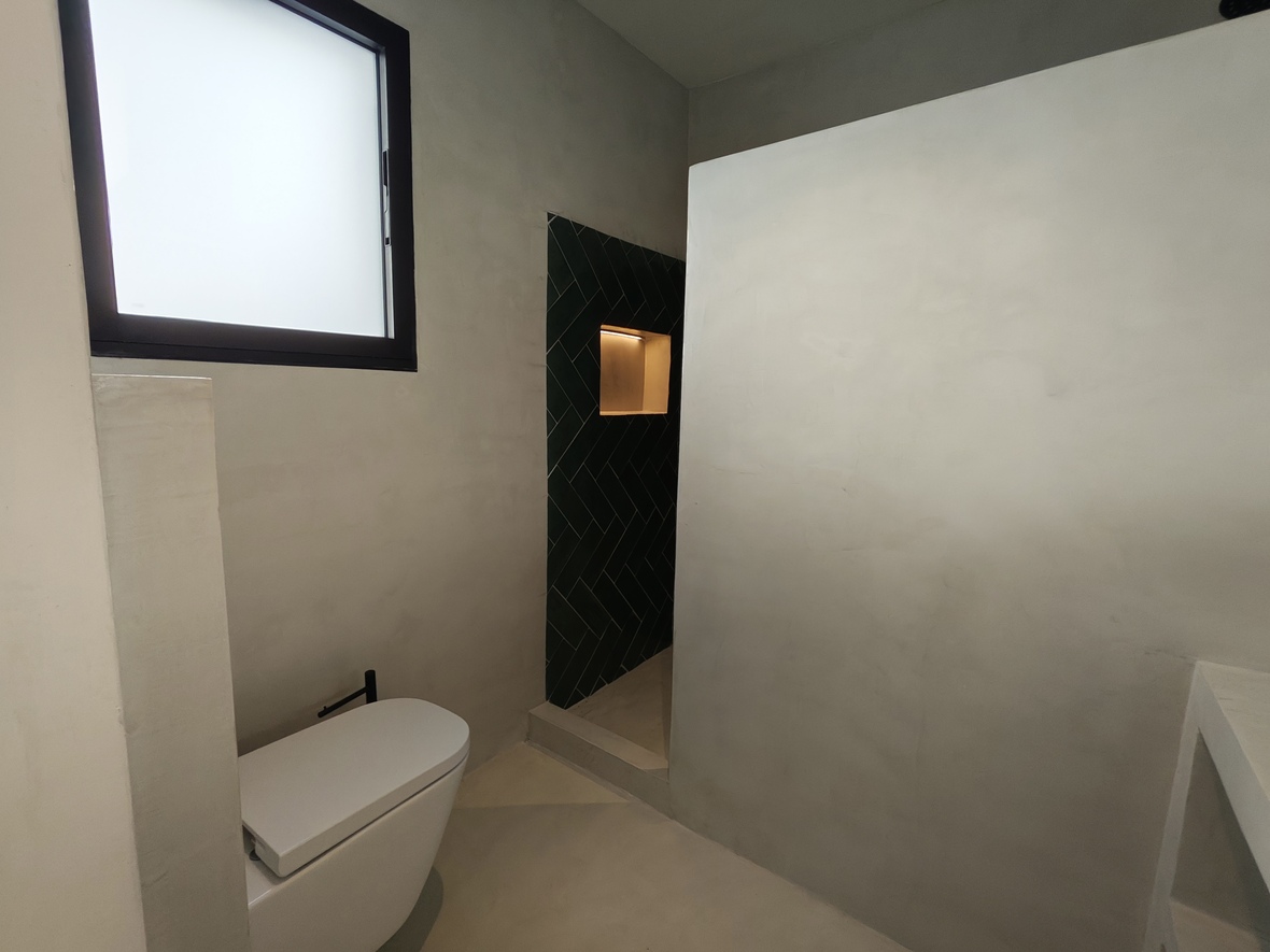 Modern bathroom featuring micro-cement walls, a wall-mounted toilet, and a walk-in shower with dark green herringbone tiles and an illuminated niche.